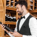 Lächelnder junger Sommelier mit Blick auf eine Flasche Wein im Weinladen