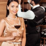 Smiling woman holding glass at wine store while steward taking bottle from shelf on background
