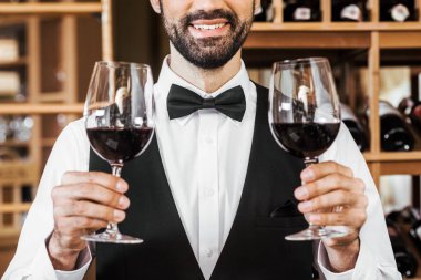 cropped shot of smiling young sommelier holding two glasses of red wine at wine store clipart