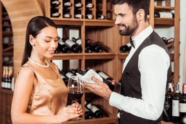 wine steward showing bottle of luxury wine to young woman at wine store clipart