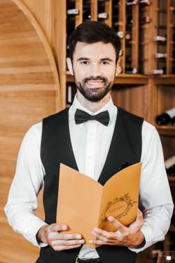 handsome young sommelier in vest and bowtie holding wine card at store clipart