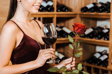 cropped shot of beautiful woman in red dress with wine and rose at wine store clipart