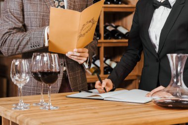 cropped shot of wine steward writing notes while customer making order at wine store clipart