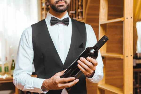 cropped shot of young wine steward with bottle of luxury wine at wine store