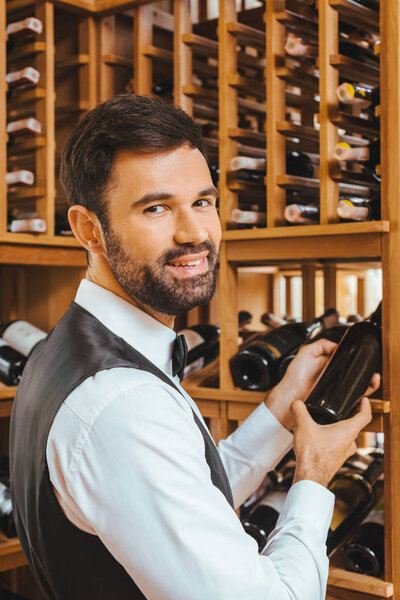 handsome young sommelier taking bottle from shelf at wine store and looking at camera