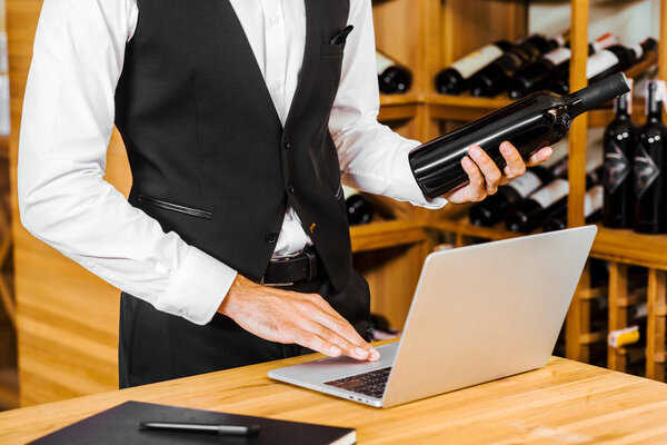 cropped shot of wine steward holding bottle and working with laptop at wine store