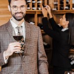 Female wine steward taking bottle from shelf for client while he looking at camera at wine store