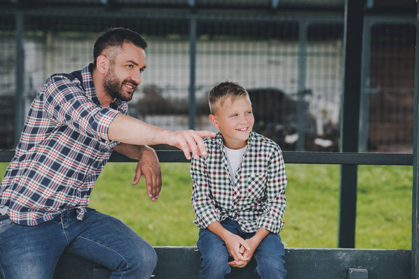 happy father and son sitting together and looking away at ranch