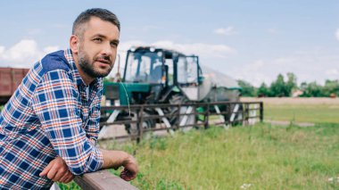 handsome middle aged farmer leaning at fence and looking away at farm  clipart