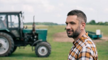 side view of handsome middle aged farmer smiling while standing near tractor in field clipart