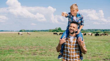 happy father carrying smiling son on neck and looking away in field clipart