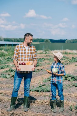happy father and son holding ripe vegetables and smiling each other in field  clipart