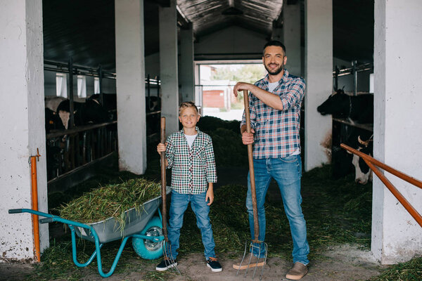 Отец и сын держат вилы и улыбаются в камеру, работая вместе в кабинке
 