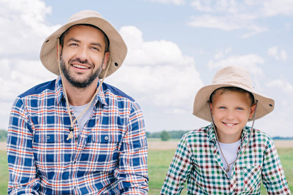 happy father and son in checkered shirts and panama hats smiling at camera outdoors