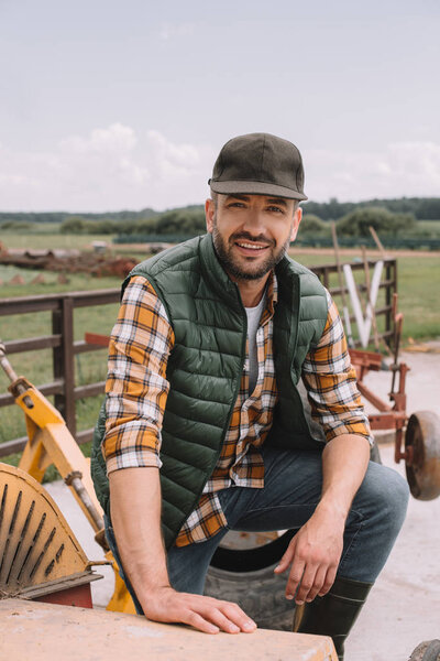 handsome middle aged farmer in cap smiling at camera while working at ranch