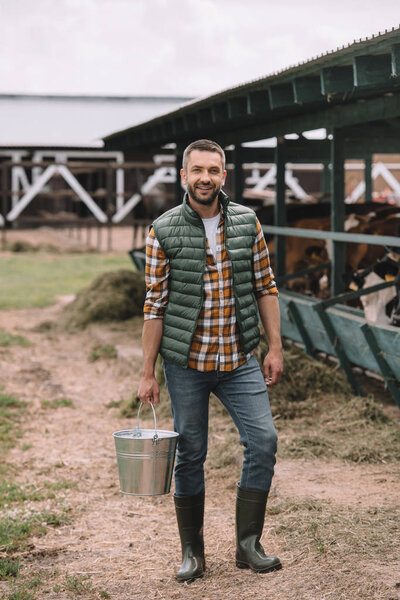handsome male farmer holding bucket and smiling at camera at cowshed