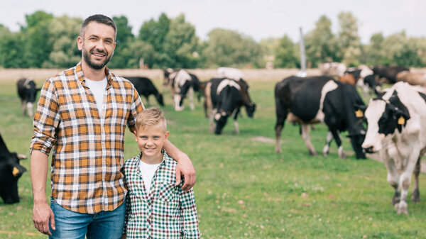 happy father and son smiling at camera while standing near grazing cattle at farm 