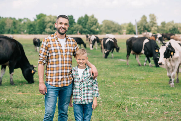 father and son smiling at camera while standing near grazing cattle at farm 