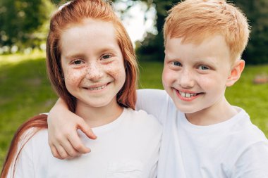 beautiful happy redhead siblings embracing and smiling at camera in park clipart