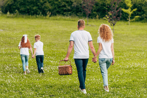 back view of family holding hands and walking with picnic basket in park