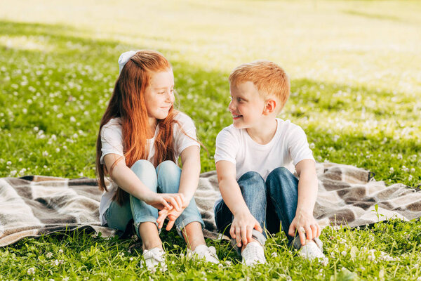 beautiful happy brother and sister smiling each other while sitting on plaid at picnic in park