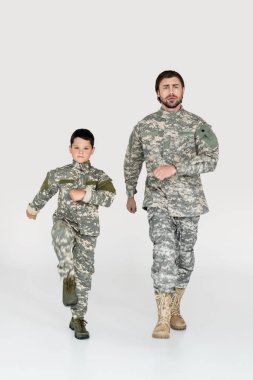 father and son in military uniforms marching and looking at camera on grey background clipart