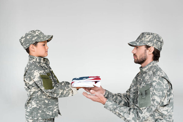 Вид сбоку маленького мальчика в камуфляжной одежде, который протягивает солдату складной американский флаг на сером фоне
