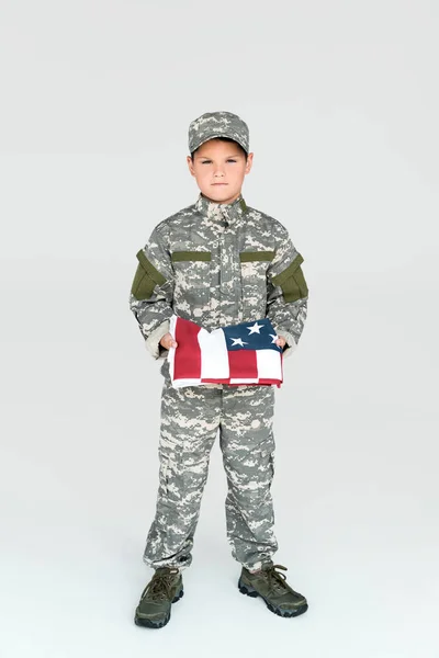 boy in military uniform holding folded american flag in hands on grey background