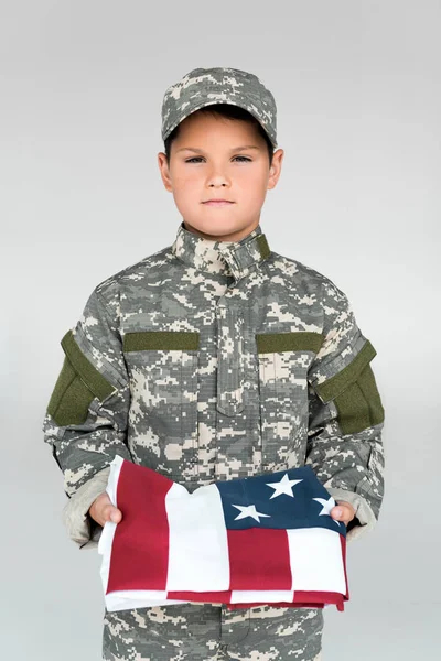 portrait of kid in military uniform holding folded american flag isolated on grey