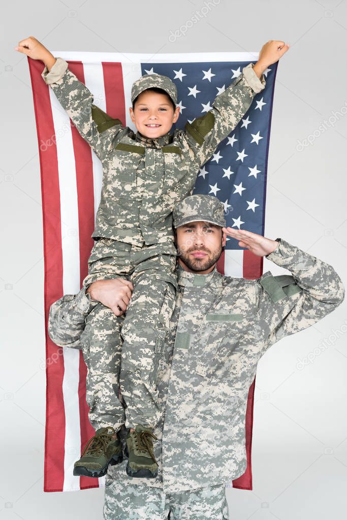 portrait of smiling boy with american flag sitting on fathers shoulder in military uniform on grey background