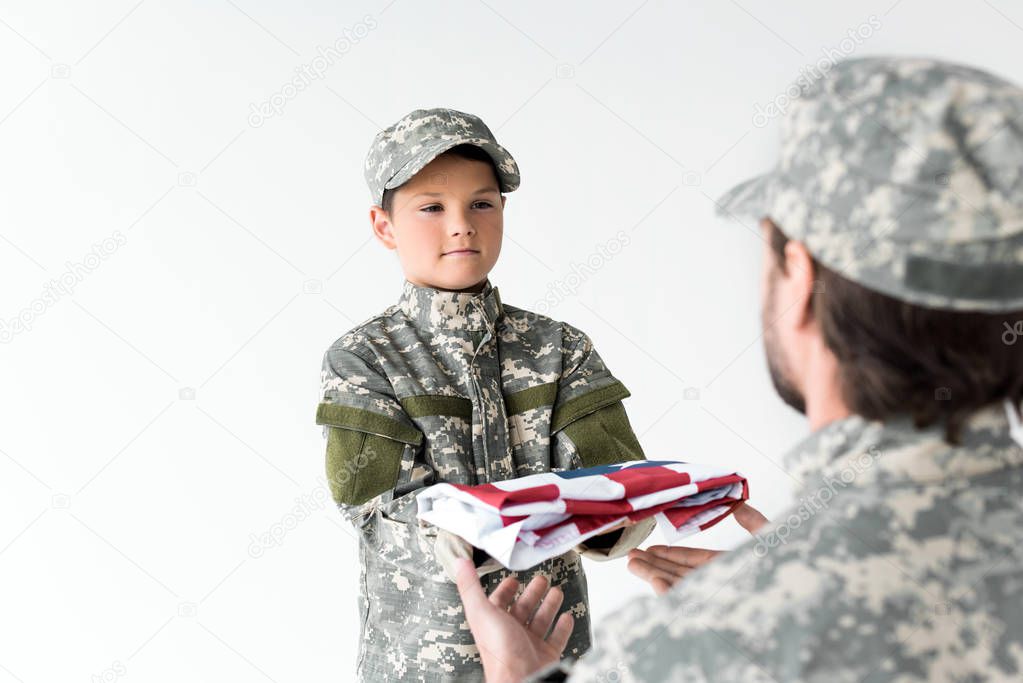 partial view of little boy in camouflage clothing giving folded american flag to soldier on grey background