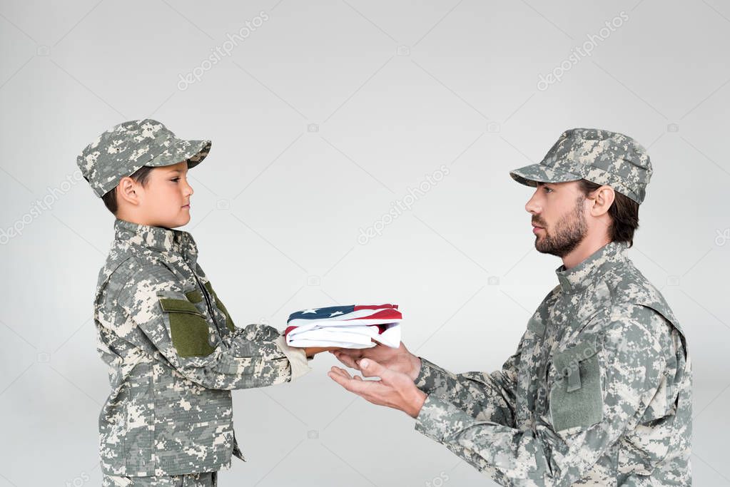 side view of little boy in camouflage clothing giving folded american flag to soldier on grey background