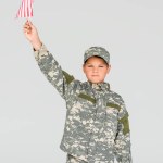 Portrait of little boy in camouflage clothing holding american flagpole in hand isolated on grey