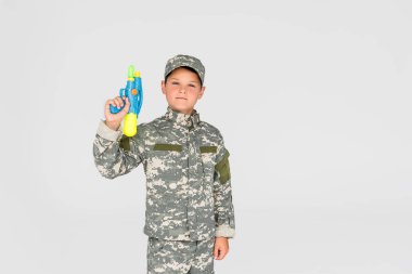portrait of boy in camouflage clothing with toy water gun isolated on grey clipart