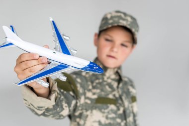 selective focus of little boy in military uniform with toy plane in hand isolated on grey clipart