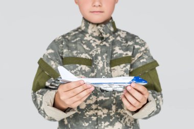 partial view of little boy in military uniform with toy plane in hands isolated on grey clipart