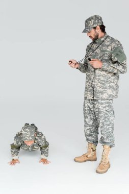 soldier in military uniform with timer and whistle controlling time while son doing push ups on grey background clipart