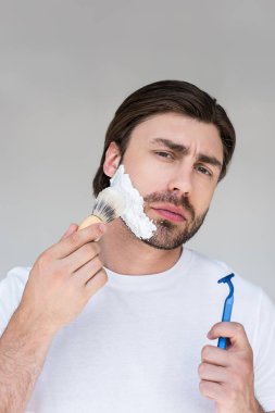 portrait of man with brush and razor in hands putting shaving foam on face on grey backdrop clipart