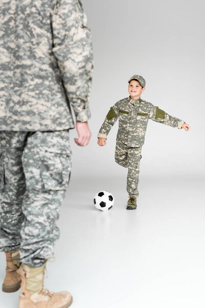 partial view of father and son in military uniforms playing soccer on grey background