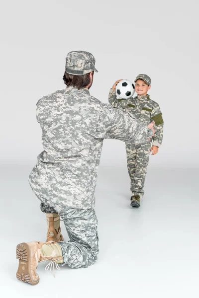 partial view of soldier with outstretched arms and smiling kid in camouflage clothing with soccer ball on grey background