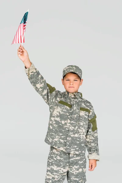 portrait of little boy in camouflage clothing holding american flagpole in hand isolated on grey