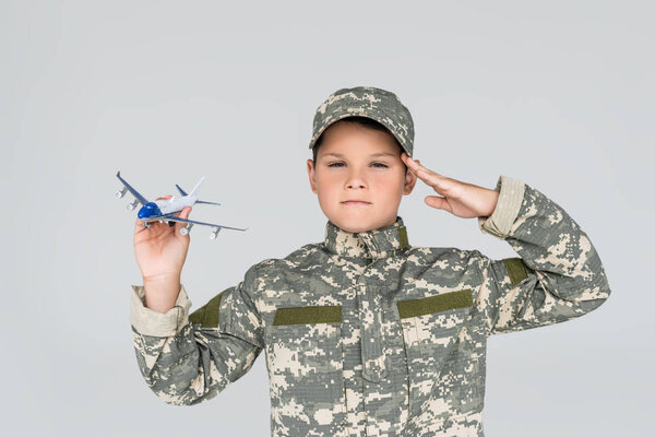 portrait of little boy in military uniform with toy plane in hand saluting isolated on grey
