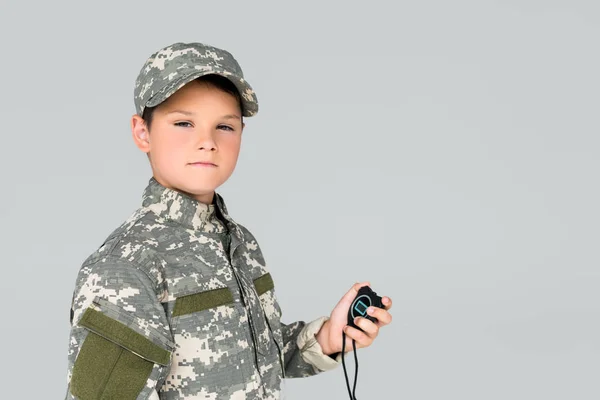 portrait of kid in military uniform with stop watch in hand looking at camera isolated on grey