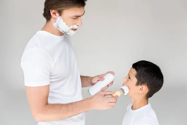 父亲的侧面在灰色背景上用刷子把剃须泡沫放在儿子脸上 — 图库照片