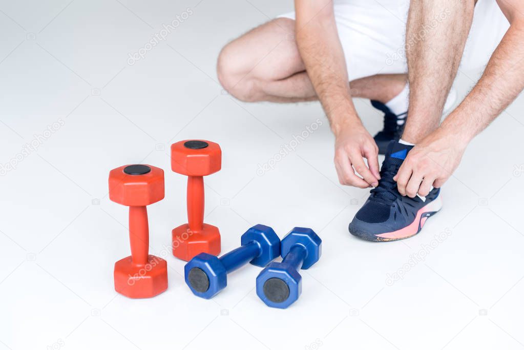 partial view of man tying shoelaces near red and blue dumbbells on grey backdrop