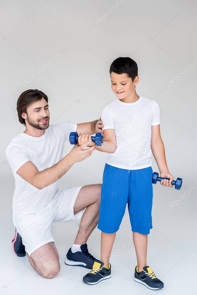 father helping little son with dumbbells to exercise on grey background