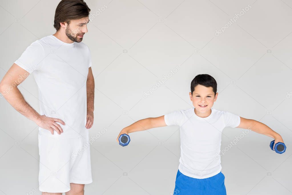 man standing akimbo and looking at son with dumbbells exercising on grey backdrop