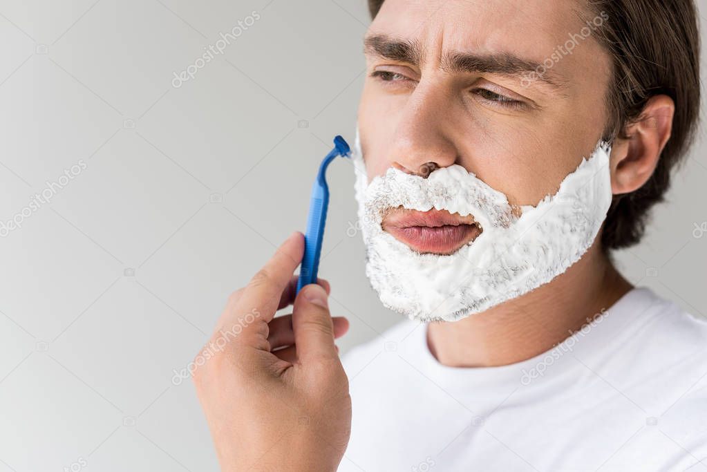 portrait of attractive man with foam on face and razor shaving beard isolated on grey