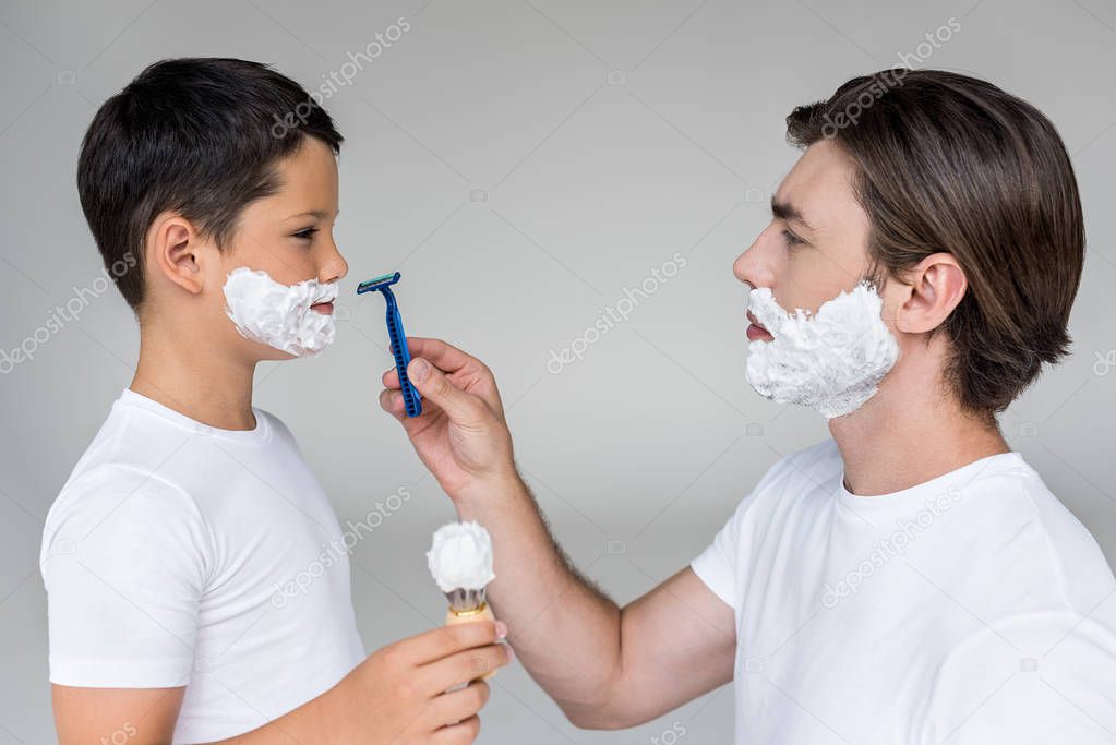 side view of father with razor in hand and son with shaving foam on faces on grey background