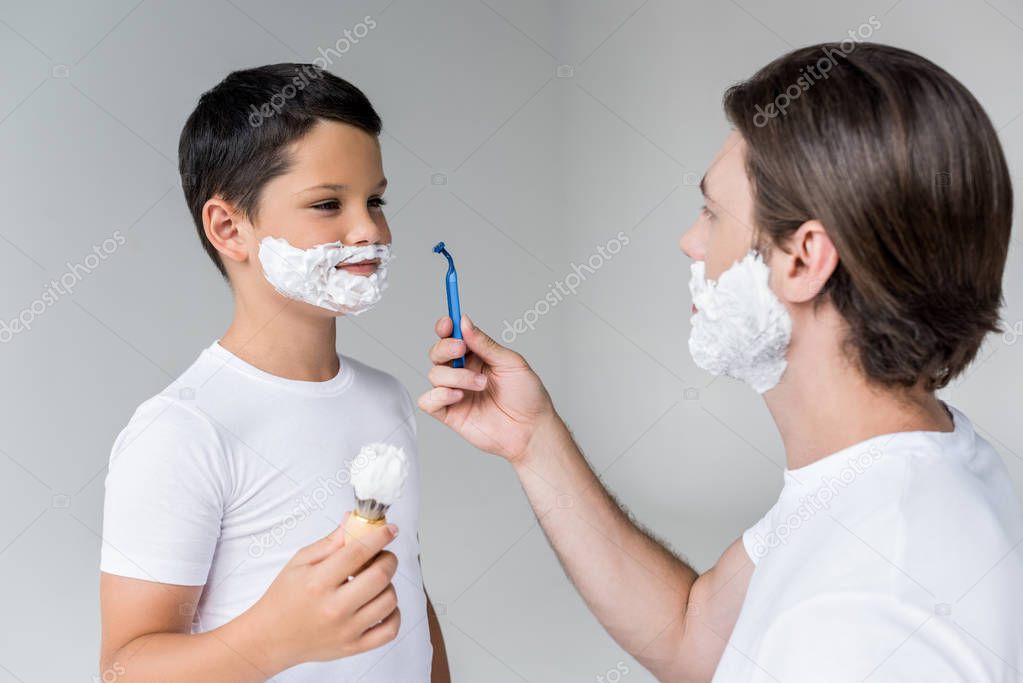 father and son with shaving foam on faces with brush and razor in hands isolated on grey